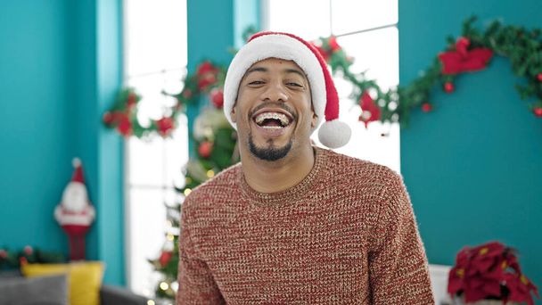 クリスマスの帽子をかぶったアフリカ系アメリカ人男性が自宅で大笑い - 写真・画像