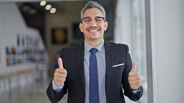 Młody Latynos mężczyzna biznes pracownik uśmiechnięty pewny siebie robi kciuki w górę gest w biurze - Zdjęcie, obraz