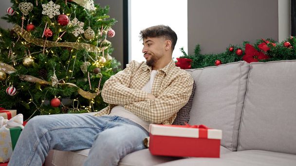 Young arab man unpacking gift looking upset sitting by christmas tree at home - Photo, Image