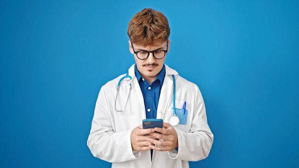 Joven médico hispano usando smartphone con cara seria sobre fondo azul aislado - Foto, imagen
