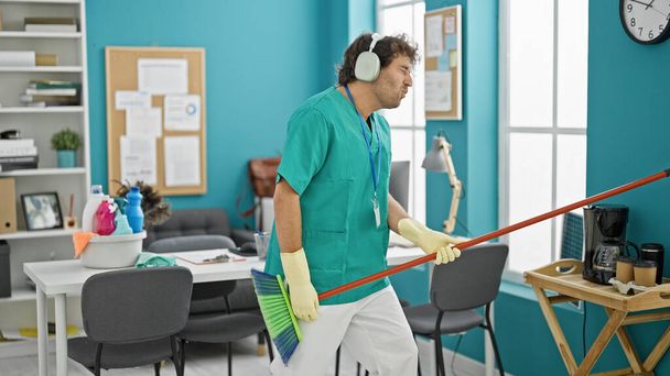 Young hispanic man professional cleaner playing air guitar dancing at office - Photo, Image