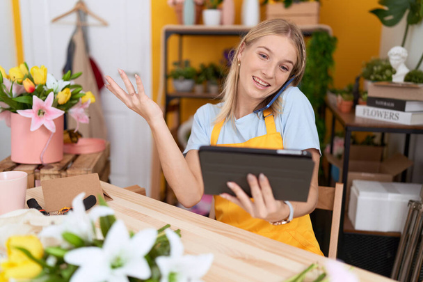 Young blonde woman florist talking on smartphone using touchpad at flower shop - Photo, image