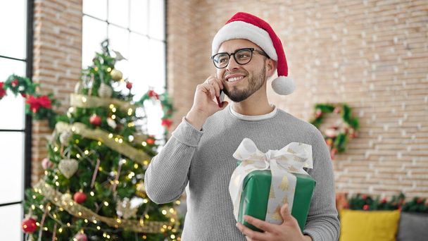 Hispanic man wearing christmas hat holding gift speaking on the phone at home - Photo, Image