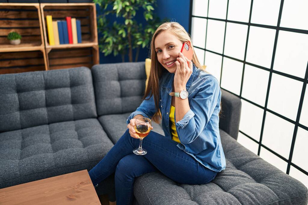 Νεαρή ξανθιά γυναίκα μιλάει στο smartphone και πίνει κρασί στο σπίτι - Φωτογραφία, εικόνα