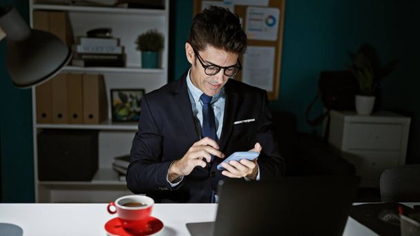 Fiatal spanyol férfi üzleti dolgozó okostelefonnal mosolyog az irodában - Fotó, kép