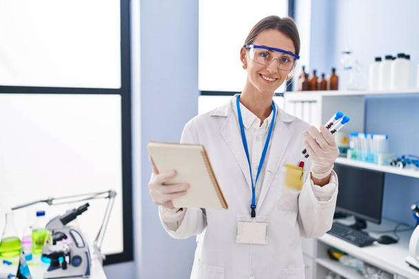 Young caucasian woman scientist holding test tubes reading notebook at laboratory - Photo, image