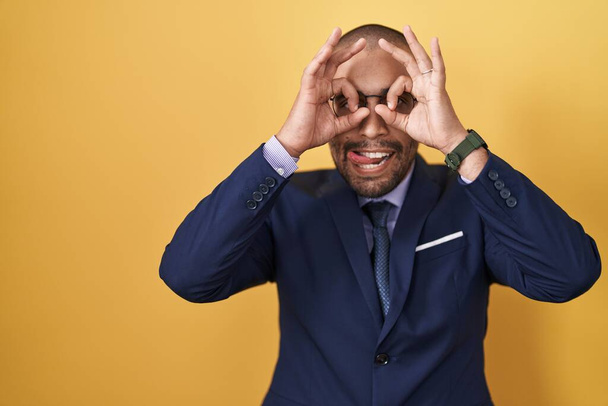 Hispanic man with beard wearing suit and tie doing ok gesture like binoculars sticking tongue out, eyes looking through fingers. crazy expression.  - Photo, Image