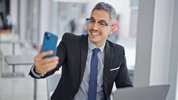 Νεαρός ισπανόφωνος επιχειρηματίας χρησιμοποιώντας φορητό υπολογιστή κάνει selfie από το smartphone στο γραφείο - Φωτογραφία, εικόνα