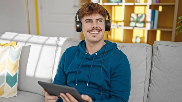 Fiatal spanyol férfi touchpad és fejhallgató ül kanapén otthon - Fotó, kép