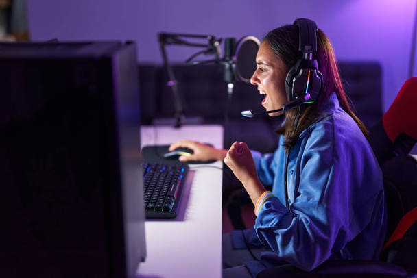 Young beautiful hispanic woman streamer playing video game with winner expression at gaming room - Photo, Image
