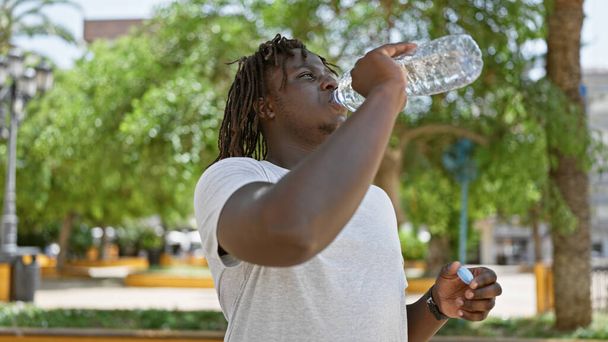 African american man standing with serious expression drinking water at park - Photo, Image