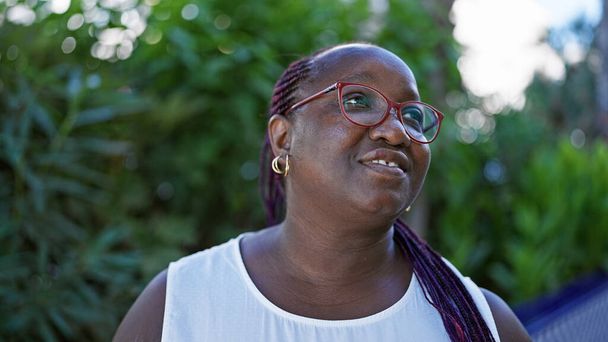 Confident african american woman with braids and glasses enjoying a day in the park, her positive expression radiating joy as she smiles looking towards the greenery and the sky. - Photo, Image