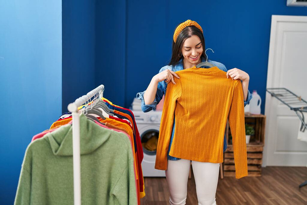 Young beautiful hispanic woman holding clothes on rack at laundry room - Foto, afbeelding
