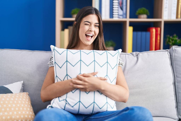Латиноамериканка обнимает подушку, сидя на диване, подмигивая, глядя в камеру с сексуальным выражением лица, веселым и счастливым лицом.  - Фото, изображение
