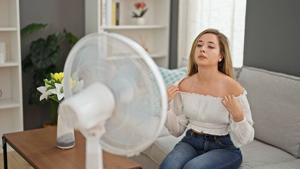 Giovane donna bionda seduta sul divano che soffre di caldo con ventilatore a casa - Foto, immagini