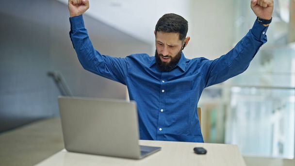 Jeune homme hispanique travailleur d'affaires en utilisant ordinateur portable et écouteurs célébrant au bureau - Photo, image