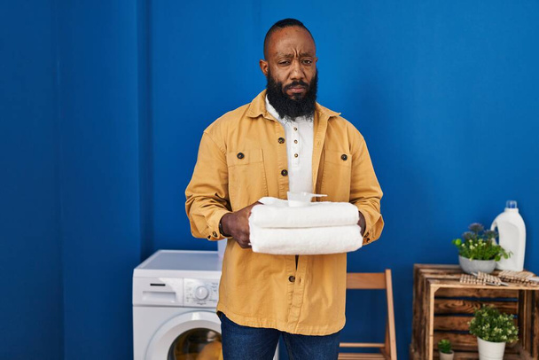 Hombre afroamericano sosteniendo toallas limpias en la lavandería escéptico y nervioso, frunciendo el ceño molesto debido a un problema. persona negativa.  - Foto, imagen