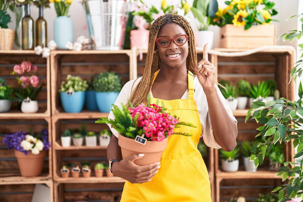 Afroamerykanka z plecionymi włosami pracująca w kwiaciarni trzymająca roślinkę uśmiechniętą pomysłem lub pytaniem wskazującym palcem szczęśliwą twarz, numer jeden  - Zdjęcie, obraz