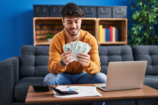 Jonge Latijns-Amerikaanse man met laptop met dollars op de bank thuis - Foto, afbeelding