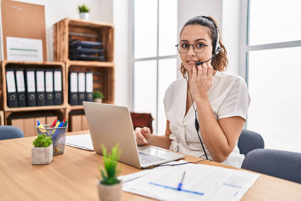 Młoda Latynoska kobieta nosząca słuchawki agenta call center w biurze wyglądająca na zestresowaną i zdenerwowaną z rękami obgryzającymi paznokcie. niepokój.  - Zdjęcie, obraz