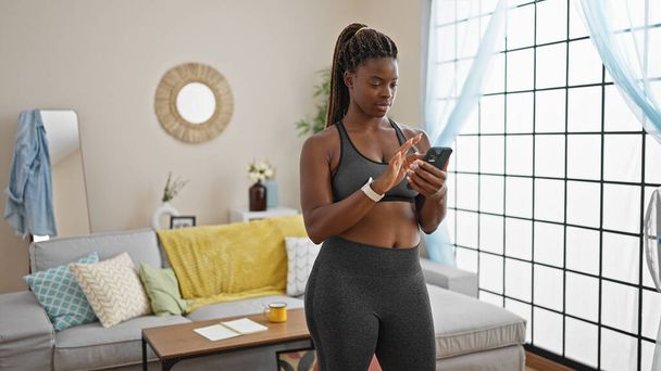 Femme afro-américaine portant des vêtements de sport en utilisant un smartphone souriant à la maison - Photo, image