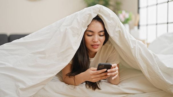 Молода красива іспаномовна жінка використовує смартфон, покритий ковдрою в спальні - Фото, зображення