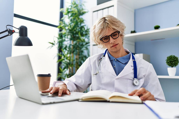 Young blond man doctor using laptop reading book at clinic - Photo, image