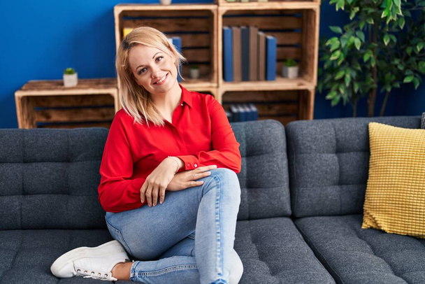 Νεαρή ξανθιά γυναίκα χαμογελά αυτοπεποίθηση κάθεται στον καναπέ στο σπίτι - Φωτογραφία, εικόνα