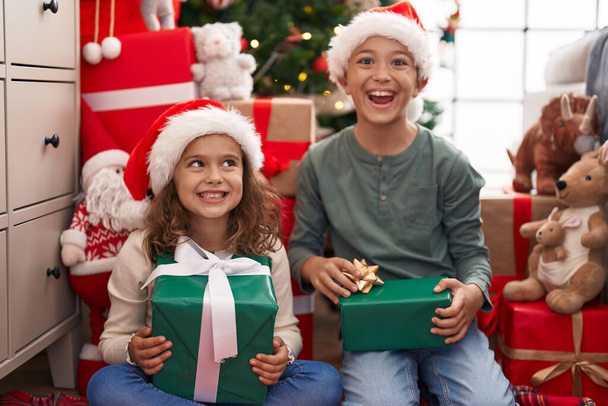 Dos niños sosteniendo regalo sentado en el suelo por el árbol de Navidad en casa - Foto, Imagen