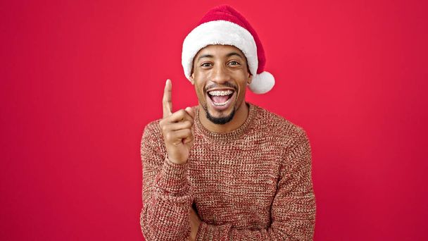クリスマスの帽子をかぶっているアフリカ系アメリカ人男性は,孤立した赤い背景の上にアイデアを持っています - 写真・画像
