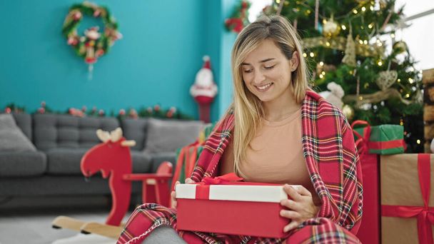 Junge blonde Frau beim Auspacken von Weihnachtsgeschenk sitzt zu Hause auf dem Fußboden - Foto, Bild
