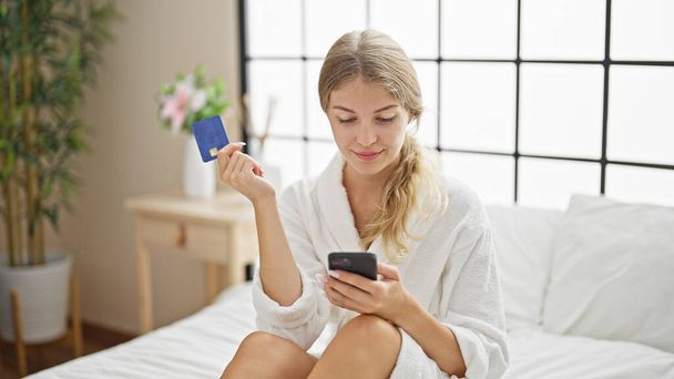 Junge blonde Frau im Bademantel mit Kreditkarte und Smartphone im Schlafzimmer - Foto, Bild