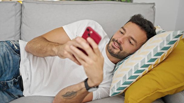 Evdeki kanepede uzanmış akıllı telefon kullanan genç bir İspanyol. - Fotoğraf, Görsel