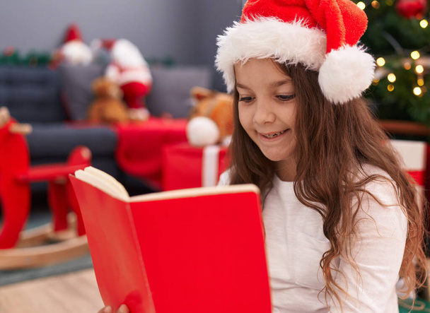 Imádnivaló spanyol lány olvasás könyvet ül a padlón karácsonyfa otthon - Fotó, kép
