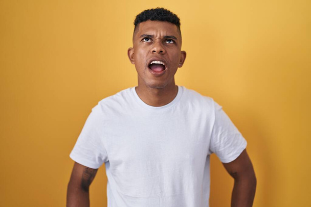 Young hispanic man standing over yellow background angry and mad screaming frustrated and furious, shouting with anger. rage and aggressive concept.  - Photo, Image