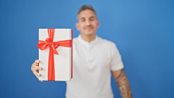 Молодой латиноамериканец улыбается уверенно держа подарок на изолированном синем фоне - Фото, изображение