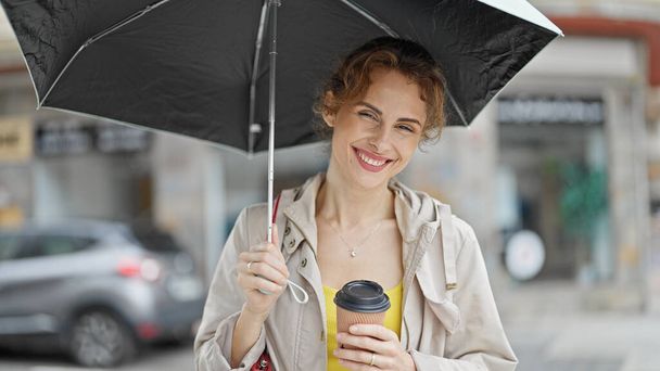 Fiatal nő kezében esernyő és kávé mosolyog az utcán - Fotó, kép