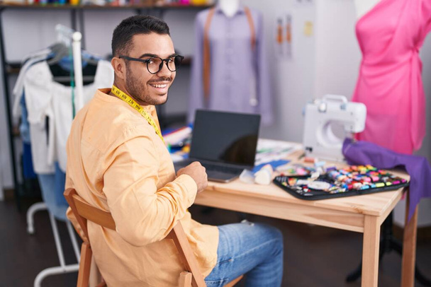 Jonge Spaanse man lachend zelfverzekerd met laptop in de kleermakerij - Foto, afbeelding