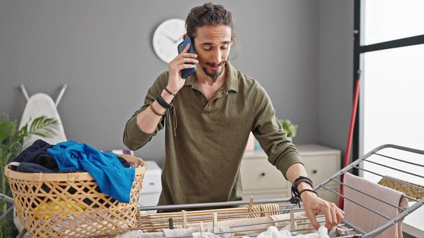 Молодий іспаномовний чоловік вішає одяг на лінії одягу, розмовляючи на смартфоні в пральні - Фото, зображення