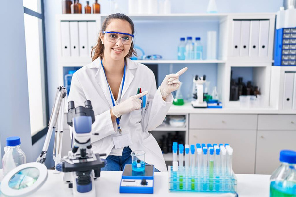 Joven mujer hispana trabajando en laboratorio científico sonriendo y mirando a la cámara apuntando con dos manos y dedos hacia un lado.  - Foto, Imagen