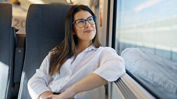 Νεαρή όμορφη ισπανόφωνη γυναίκα χαμογελά ευτυχισμένη κοιτάζοντας μέσα από το παράθυρο του τρένου βαγόνι - Φωτογραφία, εικόνα