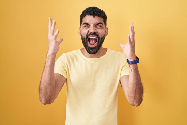 Hombre hispano con barba de pie sobre fondo amarillo celebrando loco y loco por el éxito con los brazos levantados y los ojos cerrados gritando emocionado. concepto ganador  - Foto, imagen