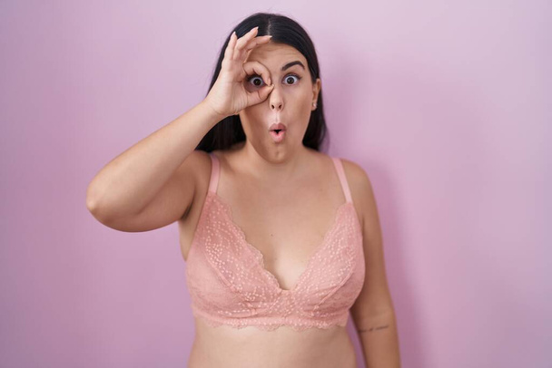 Молода іспаномовна жінка в рожевому бюстгальтері робить нормальний жест шокований здивованим обличчям, дивлячись крізь пальці. невіруючий вираз.  - Фото, зображення