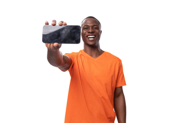 ein junger amerikanischer Mann in einem orangefarbenen Firmen-T-Shirt zeigt ein Smartphone horizontal mit einer Werbeattrappe. - Foto, Bild