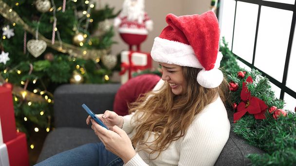 Jonge mooie Spaanse vrouw met behulp van smartphone en creditcard zitten op de bank door kerstboom thuis - Foto, afbeelding