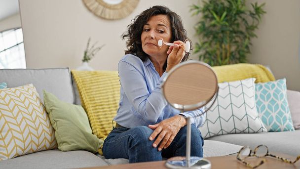 Mujer hispana de mediana edad masajeando la cara con rodillo de piel sentado en el sofá mirando en el espejo en casa - Foto, Imagen