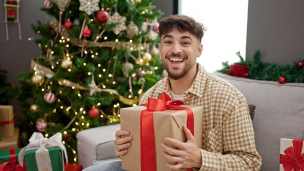 Genç Arap adam elinde hediyeyle evde Noel ağacının yanında oturuyor. - Fotoğraf, Görsel