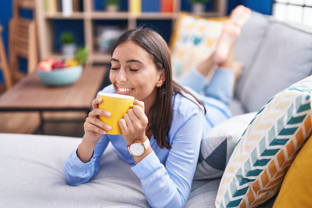 Young beautiful hispanic woman drinking coffee lying on sofa at home - 写真・画像