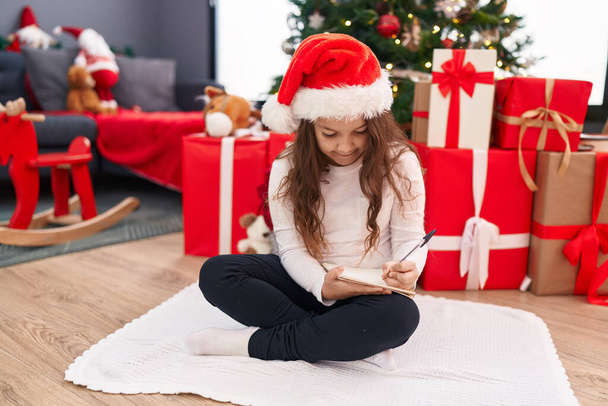 Schattige Spaanse meisje schrijven kerstman brief zitten op de vloer door kerstboom thuis - Foto, afbeelding