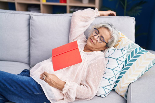 Mujer de pelo gris de mediana edad sosteniendo libro acostado en el sofá durmiendo en casa - Foto, imagen